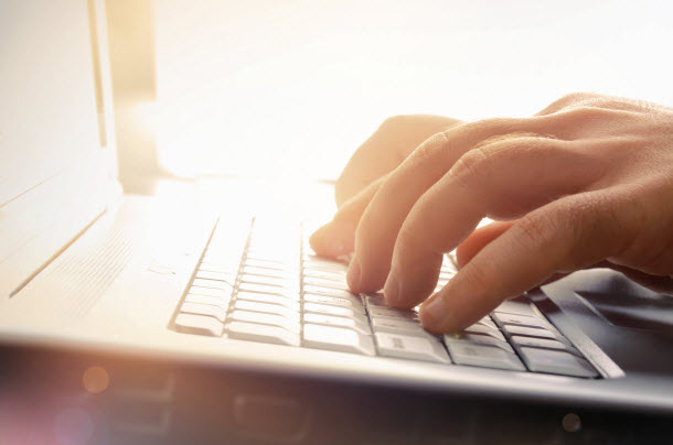 Hands typing on a laptop keyboard, with sunlight streaming in from the left, illuminating the keys.