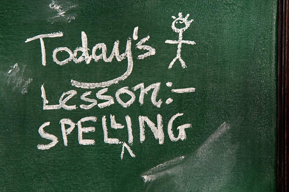 A green chalkboard with "Today's Lesson: Spelling" written in white chalk. A stick figure drawing with a crown is above the text. Smudges and chalk marks are visible on the board.