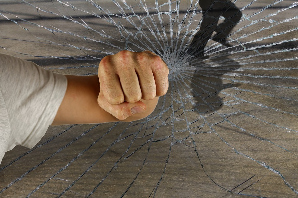 A clenched fist punching through a glass surface, causing cracks to radiate outward. A silhouetted figure is visible in the background, seen through the broken glass.