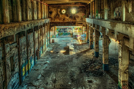 A large, abandoned industrial building with tall concrete pillars and walls covered in colorful graffiti. Sunlight filters through broken windows, illuminating the dusty and debris-littered floor.