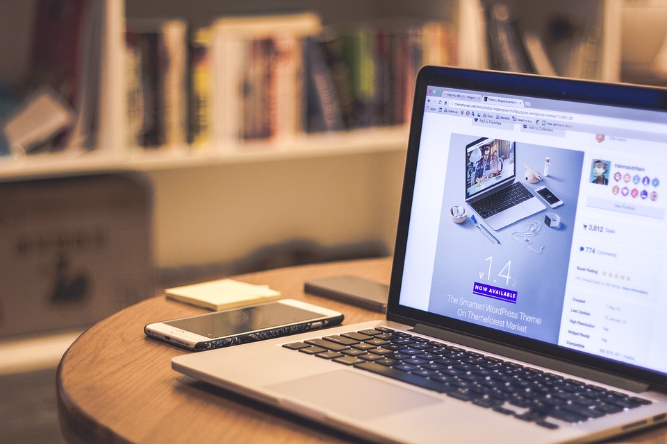 A laptop displaying a website is on a wooden table. Next to it are a smartphone and a pen. In the background, there is a blurred bookshelf filled with various books. The setting appears to be a cozy home office or study area.