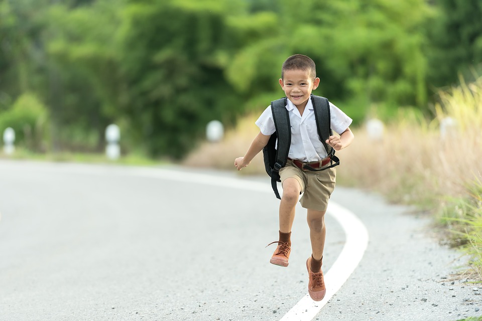 A young boy wearing a backpack, white shirt, and khaki shorts is joyfully running on a paved road with greenery in the background.