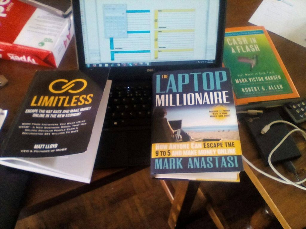 A cluttered desk with a laptop displaying a spreadsheet. Surrounding it are books titled "Limitless," "The Laptop Millionaire," and "Cash in a Flash." Cables and a phone are also visible on the desk.
