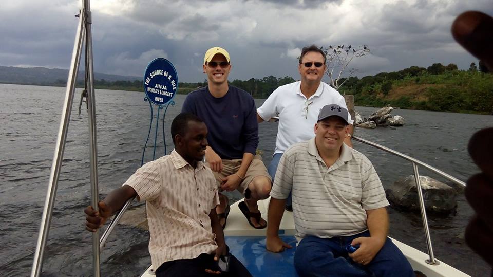 Four men are on a boat near a sign for Jinja Nile Explorers Club in a lake. One is wearing a hat and sunglasses. They're smiling with a cloudy sky and trees in the background.