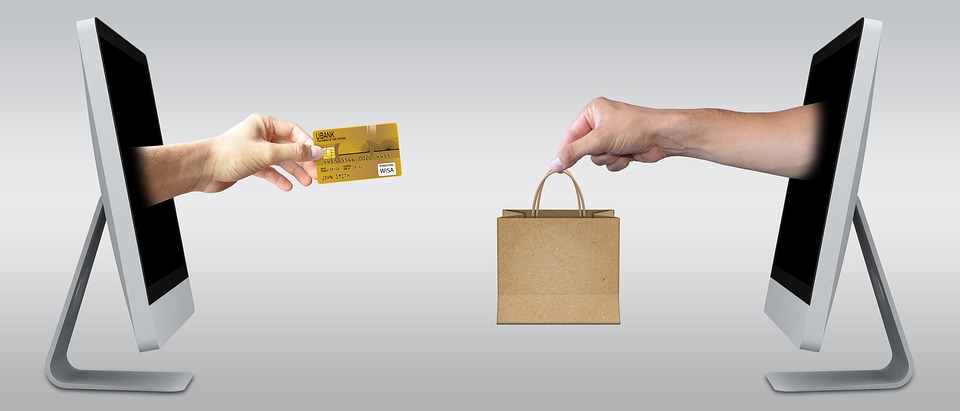 Two computer screens facing each other display hands reaching out. One hand holds a credit card, while the other holds a shopping bag, symbolizing online shopping. The background is a simple gradient.