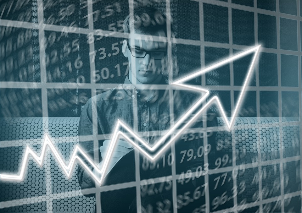 A person wearing glasses and a button-up shirt looks at a tablet, standing in front of a grid-like background with a stylized upward-pointing arrow and various numbers, symbolizing financial growth and analysis.