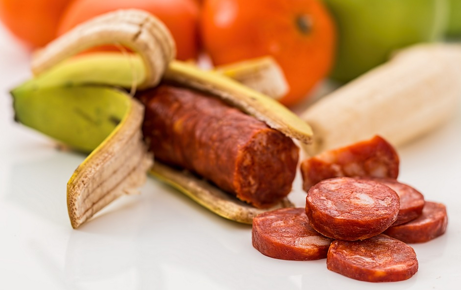 A peeled banana with a sausage replacing the fruit is on a white surface. A few slices of sausage are in front. In the blurred background, there are whole bananas and orange fruits.