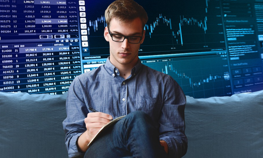 A person wearing glasses is seated and writing in a notebook. They are in front of a digital background displaying financial graphs and stock market data on multiple screens.