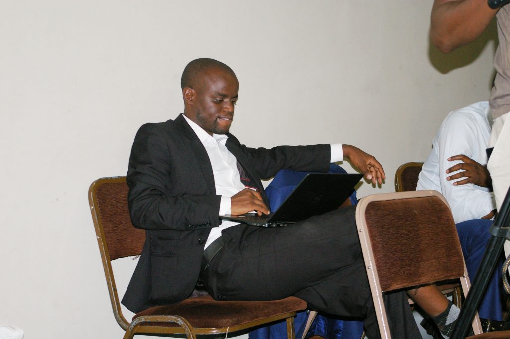A person in a black suit sits on a chair, using a laptop positioned on their lap. They appear focused, with one arm resting on the chair next to them. The background is a plain white wall.
