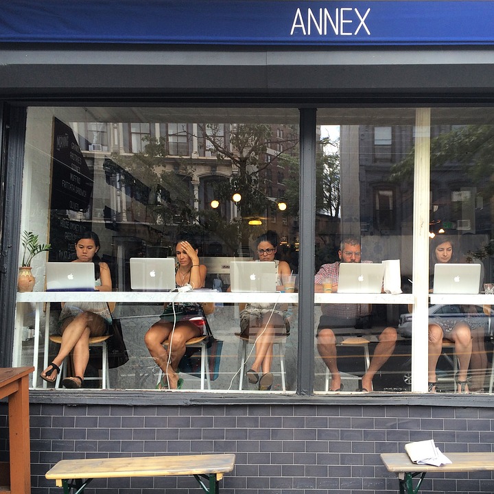 Five people sit inside a cafe, working on laptops by a large window. The cafe sign reads "ANNEX." The street and buildings outside are reflected in the window.