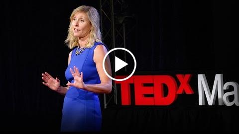 A woman in a blue dress is speaking on stage at a TEDx event. She is gesturing with her hands and standing in front of a black background with "TEDx" in red and white beside her.