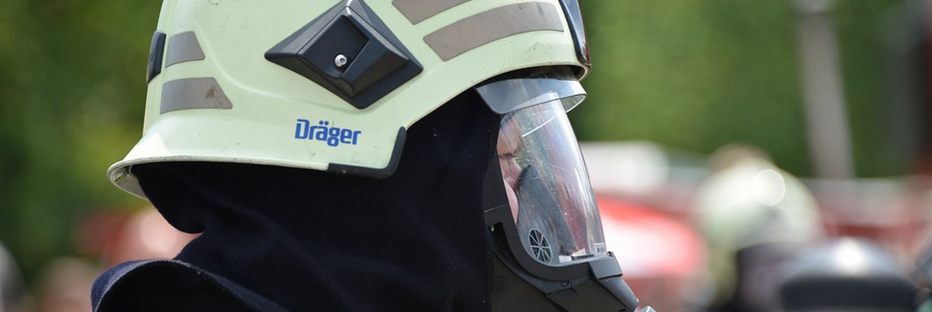 A firefighter wearing a beige helmet with a clear visor and protective suit is shown in profile. A blurred outdoor background is visible.