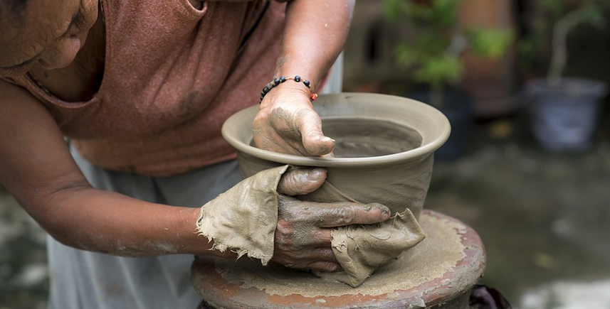 A person shaping a clay pot on a pottery wheel. Their hands are covered in clay, and they're using a cloth for support. The background is slightly blurred, with greenery visible.