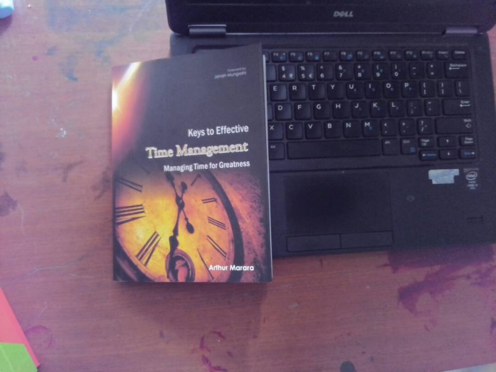 A book titled "Keys to Effective Time Management: Managing Time for Greatness" by Arthur Marara is placed on a Dell laptop keyboard. The book cover features a clock face. The laptop is on a wooden surface.