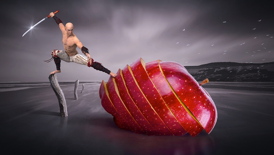 A martial artist poses dramatically with a sword on a beach, standing on a wooden post. In front of him is a large, sliced apple, creating a surreal scene. The sky is overcast, and the background is a vast, empty shoreline.