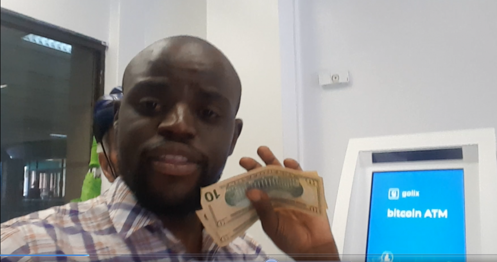 A person is standing indoors, holding a ten-dollar bill and another currency note. A Bitcoin ATM is visible in the background. They are wearing a plaid shirt and looking at the camera.