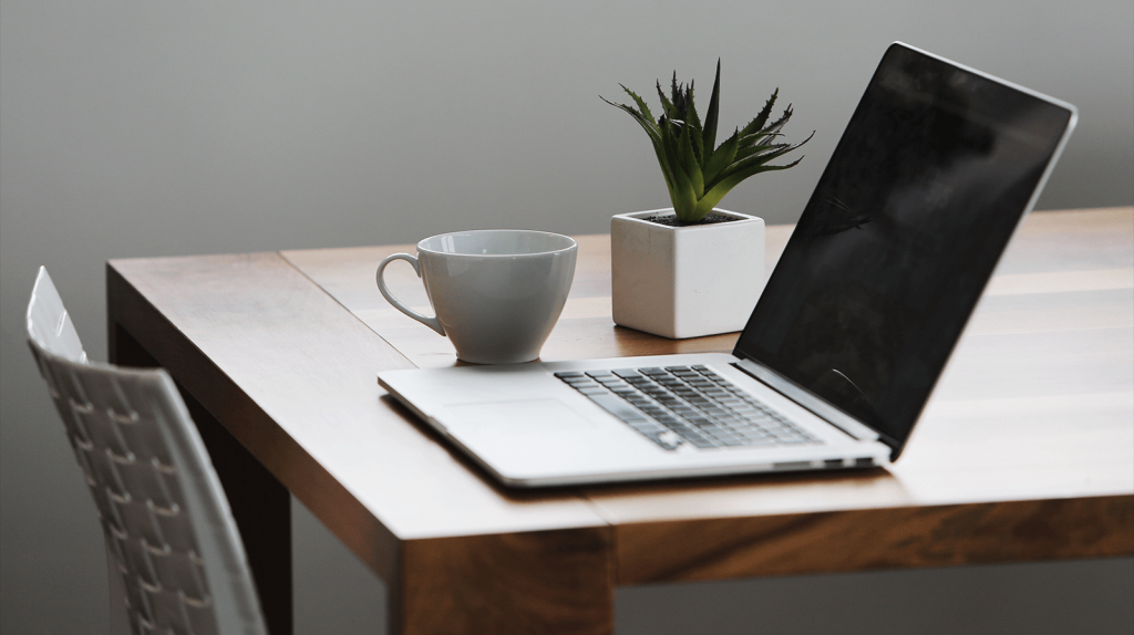 A wooden desk with a silver laptop, a white coffee cup, and a small potted plant. The laptop is open, and the cup is empty. The plant is in a white square pot. A white chair is partially visible beside the desk.