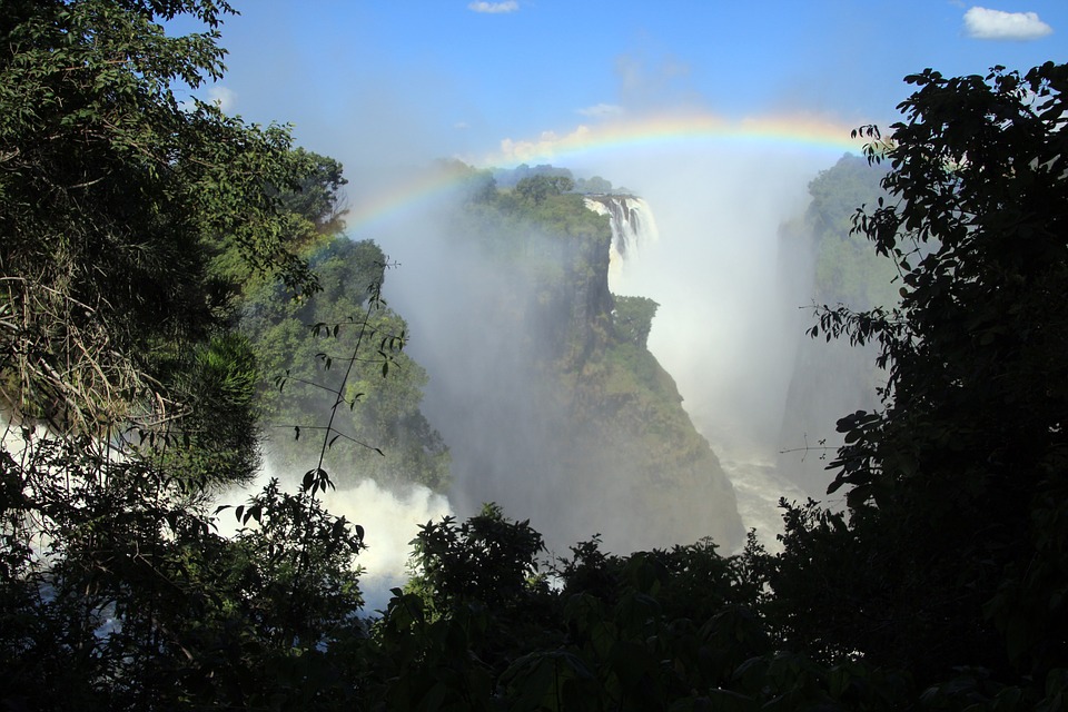 A wide waterfall cascades into a gorge surrounded by lush greenery. Mist rises from the water, and a vivid rainbow arches across the clear blue sky.