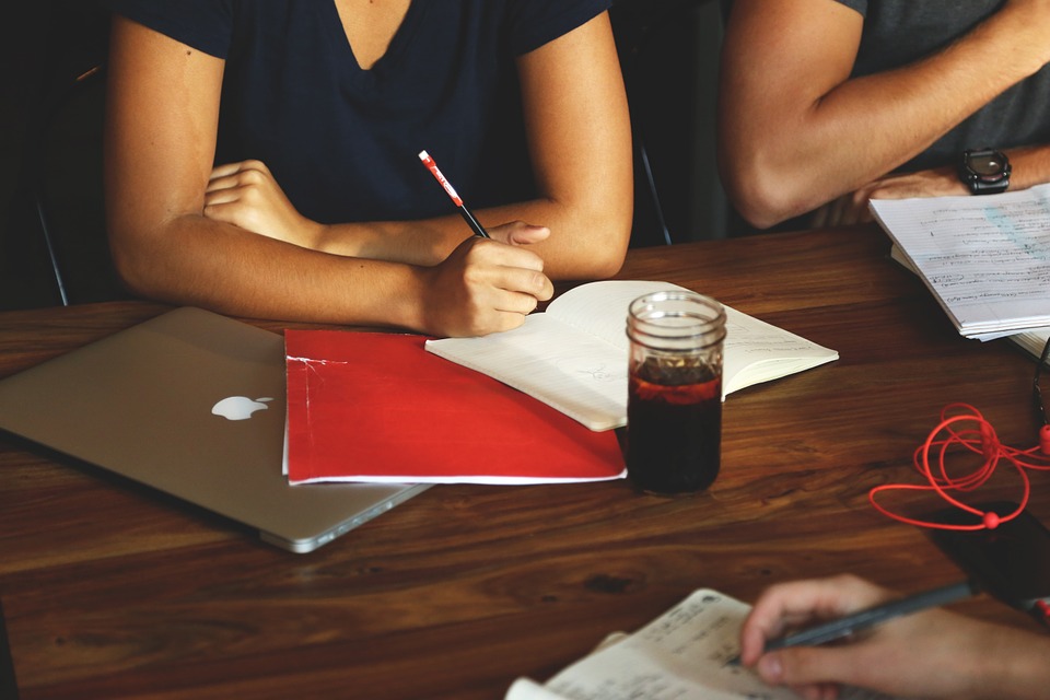 Two people sit at a wooden table, one writing in a notebook with a red pencil next to a red folder and a laptop. A glass of dark liquid with a lid and a notebook with notes are also on the table.