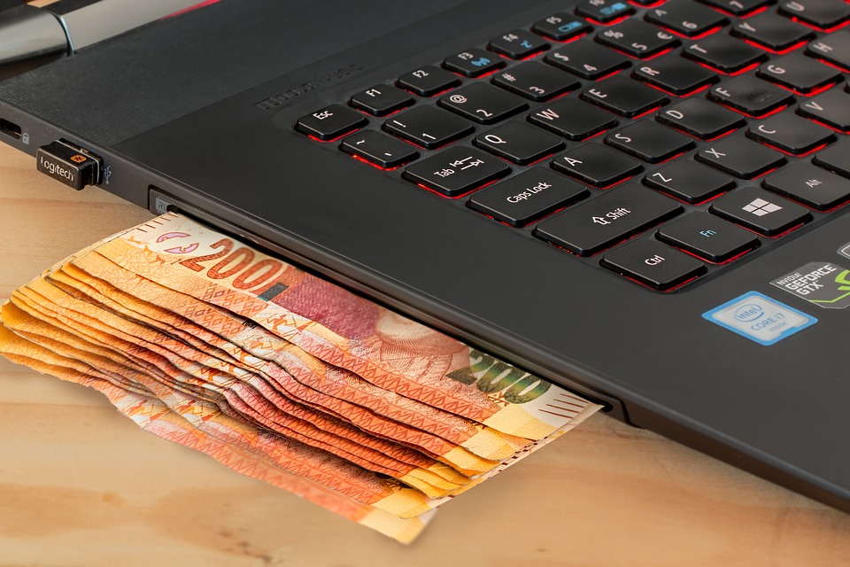 A laptop with euro banknotes partially sticking out from the CD/DVD drive, resting on a wooden surface.