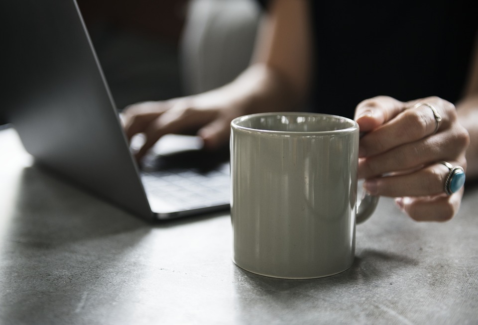 A person is using a laptop while holding a gray mug. Their hand rests on a table, wearing a ring with a turquoise stone. The scene suggests a casual or work-from-home setting with a focus on multitasking.