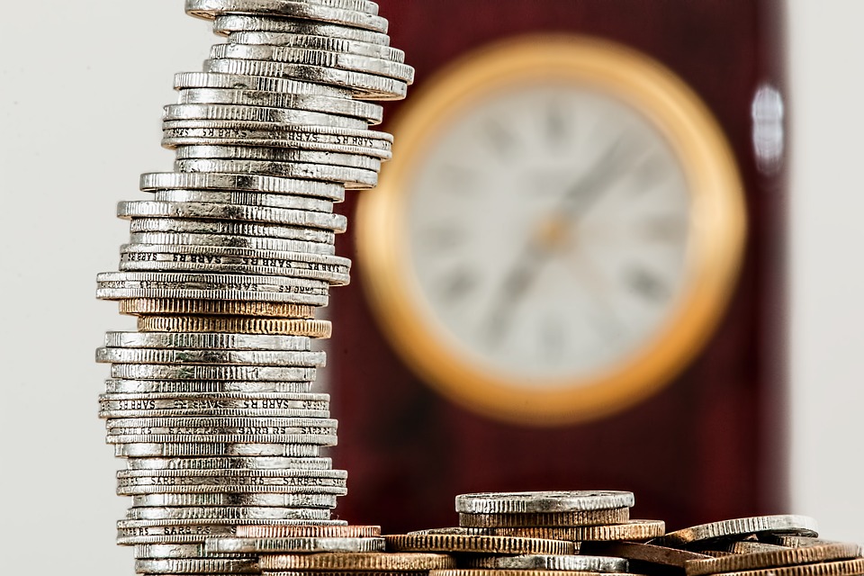 A tall stack of silver coins is precariously balanced in the foreground, with additional coins scattered below. A blurred analog clock in the background suggests a theme of time and money.