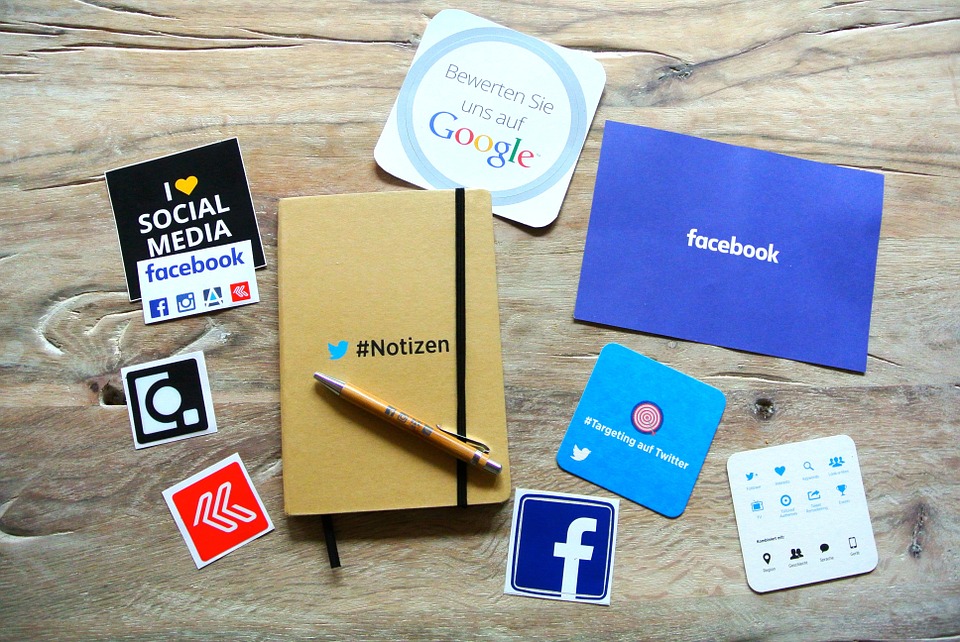 A wooden desk with a brown notebook labeled "#Notizen," surrounded by social media icons and stickers for Facebook, Instagram, Google, and Twitter. A pen rests on the notebook.