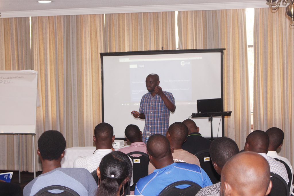 A person stands in front of a seated audience giving a presentation. A projector screen displays content behind them, and a flip chart is visible on the side. The setting appears to be a conference room with drawn curtains.