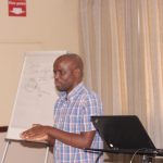A man in a checkered shirt stands in front of a flip chart, speaking next to a laptop on a podium. There is a fire exit sign on the wall behind him and a curtain to the side. The flip chart has some writing and diagrams.
