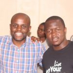 Two men are standing together indoors. One is wearing a plaid shirt, and the other a black t-shirt with white text. They are smiling and have their arms on each other's shoulders. A person in the background is slightly out of focus.