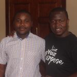 Two men are standing indoors in front of a wooden door. The man on the left is wearing a checked shirt, while the man on the right is wearing a black t-shirt with the text "Live Your Purpose." They are looking at the camera.