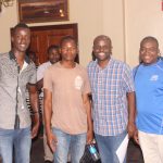 Four men standing and smiling in a warmly lit room. One holds a bottle of water. They are casually dressed, with two wearing patterned shirts, one in a brown t-shirt, and another in a blue sports shirt. Other people are visible in the background.