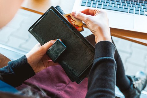 A person holding a black wallet, inserting a card into it. A laptop is visible on the table in the background. The scene suggests a casual setting, possibly in a café or a home.
