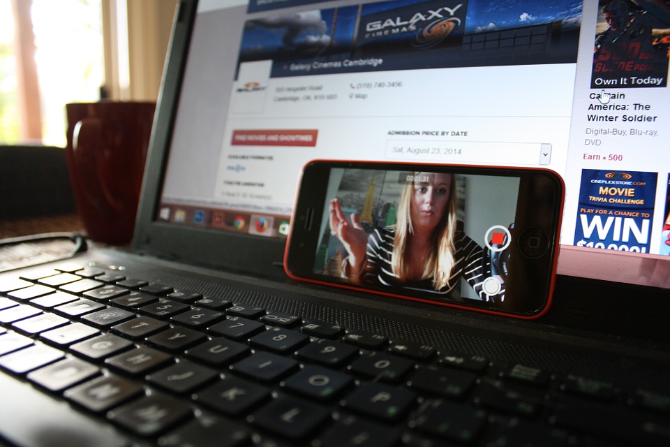 A smartphone placed on a laptop keyboard, showing a paused video of a person talking. The laptop screen displays a movie booking website. A red mug is in the background.