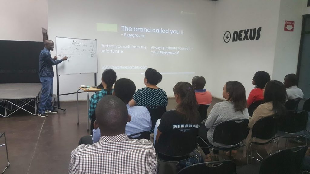 A speaker presents to an audience in a classroom setting. The slide on the screen reads "The brand called you - Playground" with additional notes about self-promotion and protection. Attendees sit in rows, attentively facing the speaker and screen.