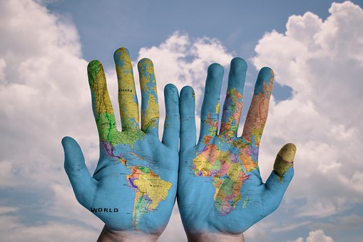Two open hands against a cloudy sky, painted with a map of the world. Each hand shows a different hemisphere, with continents and oceans visible, symbolizing global unity and diversity.
