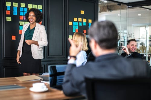 A woman stands smiling near a black wall with colorful sticky notes. She is speaking to three seated colleagues in a modern conference room. A cup and saucer are on the table in the foreground.