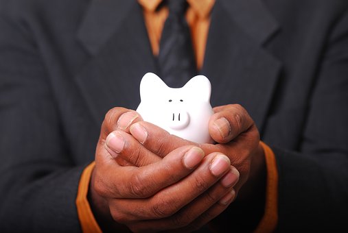 A person in a suit holds a small white piggy bank in both hands, symbolizing saving or financial security. The focus is on the piggy bank, with the individual's face not visible.