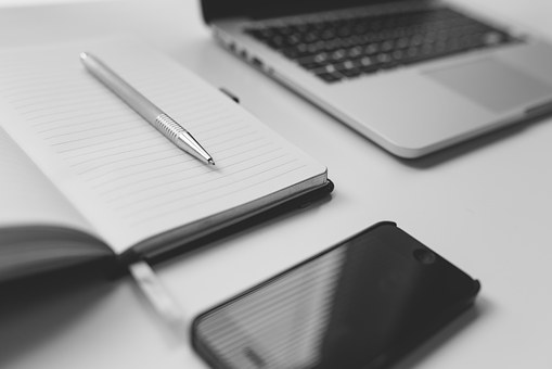Black and white image showing an open notebook with a pen resting on its pages, next to a smartphone and a laptop on a white surface.