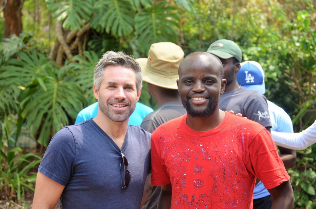 Two men smiling and standing together outdoors. The man on the left is wearing a gray t-shirt and sunglasses, while the man on the right is wearing a red t-shirt with a Bitcoin symbol. Other people and large green leaves are visible in the background.