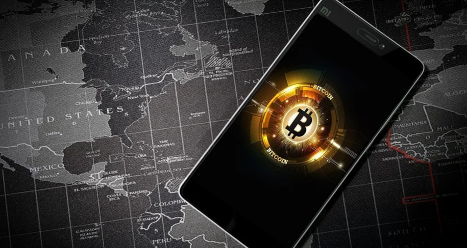 A smartphone displaying a glowing Bitcoin symbol on its screen is placed on a dark world map. The map shows parts of North and South America. The Bitcoin symbol is surrounded by digital, circuit-like patterns.