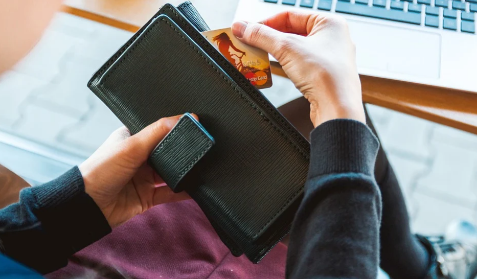 A person holding a black wallet, inserting a small card into it. A laptop is visible on the desk in the background. The person appears to be wearing a dark-colored long-sleeve shirt.