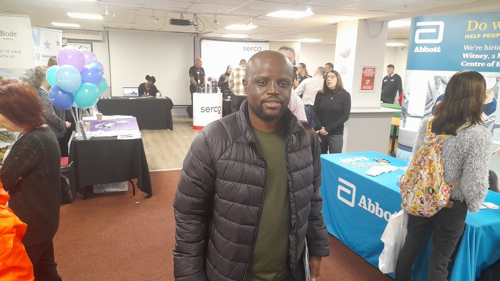 A man wearing a black jacket stands smiling in a room with various company booths and people in the background. The booths display banners and materials, and there are balloons on one side. The setting appears to be a career or job fair.