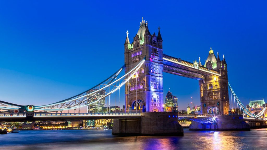 A view of Tower Bridge in London at dusk, illuminated with colorful lights, captivates those moving to the United Kingdom from Zimbabwe. The sky is a deep blue with the city skyline in the background, as the Thames River flows beneath this iconic structure.